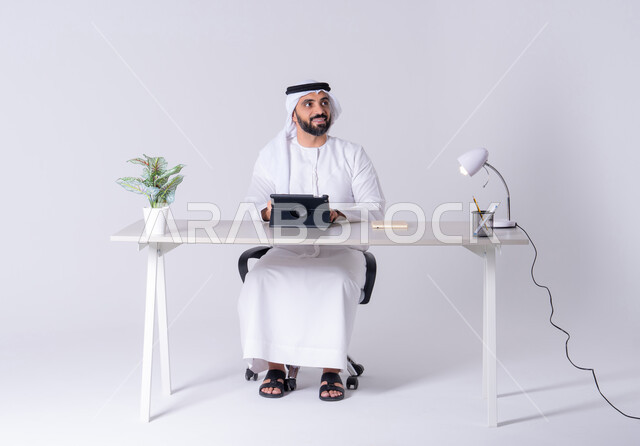 اجراء مكالمه فيديو ،دمج التقنيات المتطوره فى الحياة اليومية،انجاز مهام العمل عن بعد ،بورتريه لرجل عربي خليجي اماراتي يرتدي الكندوره والغتره يجلس عل مكتب ويمسك التابلت بيده