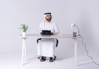 اجراء مكالمه فيديو ،دمج التقنيات المتطوره فى الحياة اليومية،انجاز مهام العمل عن بعد ،بورتريه لرجل عربي خليجي اماراتي يرتدي الكندوره والغتره يجلس عل مكتب ويمسك التابلت بيده