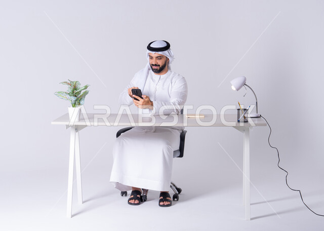 دمج التقنيات المتطوره فى الحياة اليومية، الدردشة والتحدث مع الأصدقاء، بورتريه لرجل عربي خليجي اماراتي يرتدي الكندوره والغتره يجلس عل مكتب ويمسك الهاتف المحمول بيده