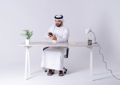دمج التقنيات المتطوره فى الحياة اليومية، الدردشة والتحدث مع الأصدقاء، بورتريه لرجل عربي خليجي اماراتي يرتدي الكندوره والغتره يجلس عل مكتب ويمسك الهاتف المحمول بيده