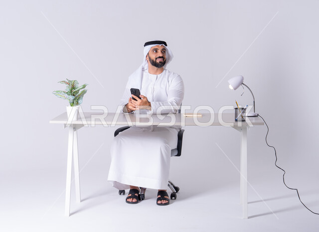 استخدام التقنيات والتكنولوجيا الحديثة بالحياة اليومية، الدردشة والتحدث مع الأصدقاء، عرض شاشه الجوال ،بورتريه لرجل عربي خليجي اماراتي يرتدي الكندوره والغتره يجلس عل مكتب ويمسك الهاتف المحمول بيده