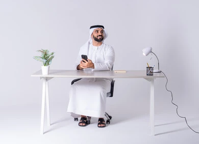 استخدام التقنيات والتكنولوجيا الحديثة بالحياة اليومية، الدردشة والتحدث مع الأصدقاء، عرض شاشه الجوال ،بورتريه لرجل عربي خليجي اماراتي يرتدي الكندوره والغتره يجلس عل مكتب ويمسك الهاتف المحمول بيده