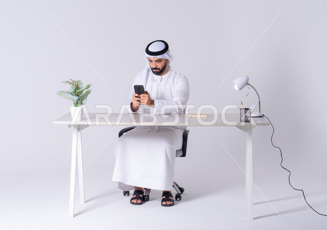 الدردشة والتحدث مع الأصدقاء، عرض شاشه الجوال ،بورتريه لرجل عربي خليجي اماراتي يرتدي الكندوره والغتره يجلس عل مكتب ويمسك الهاتف المحمول بيده، استخدام التقنيات والتكنولوجيا الحديثة بالحياة اليومية