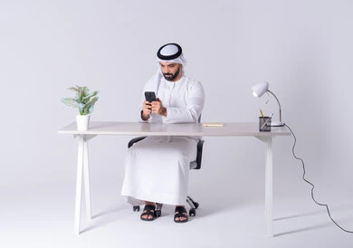 الدردشة والتحدث مع الأصدقاء، عرض شاشه الجوال ،بورتريه لرجل عربي خليجي اماراتي يرتدي الكندوره والغتره يجلس عل مكتب ويمسك الهاتف المحمول بيده، استخدام التقنيات والتكنولوجيا الحديثة بالحياة اليومية