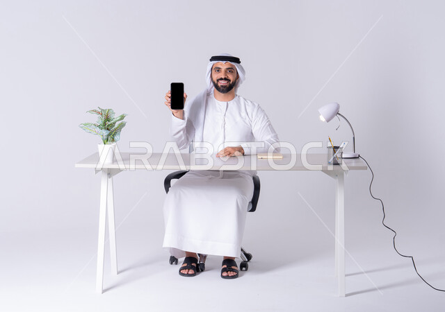 عرض شاشه الجوال ، الدردشة والتحدث مع الأصدقاء، بورتريه لرجل عربي خليجي اماراتي يرتدي الكندوره والغتره يجلس عل مكتب ويمسك الهاتف المحمول بيده، استخدام التقنيات والتكنولوجيا الحديثة بالحياة اليومية