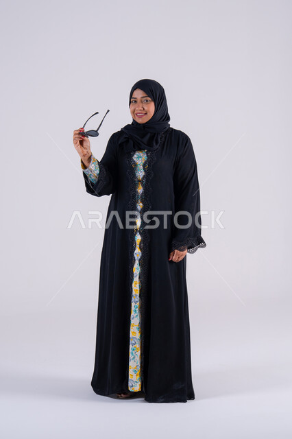 A Saudi woman wearing black sunglasses with an expression of pleasure and happiness, a concern for elegance and attention to one's outward appearance, the latest fashion trends, wearing sunglasses, a portrait of an Arab Gulf Emirati woman wearing an abaya and hijab, standing with crossed hands and gestures of self-confidence.