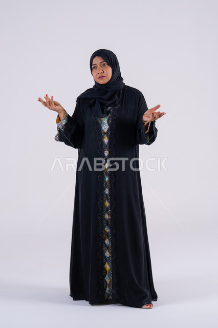 Trying to find a solution to difficult problems, anxiety and confusion in making the right decision, deep thinking and absent-mindedness, a portrait of an Arab Gulf Emirati woman wearing an abaya and hijab, raising her hand, a Saudi woman standing with gestures of integration.