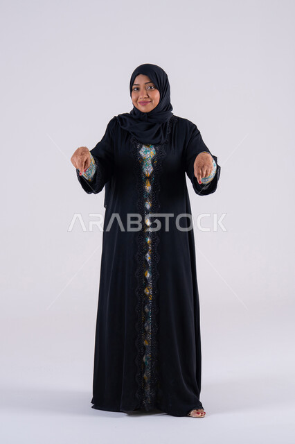 تعابير تحدد هذا المكان هنا، سيده سعوديه تحدد موقع معين ، بورتريه لامراه عربيه خليجيه اماراتيه ترتدي العباءه والحجاب تشير بإصبع السبابة لاسفل إلى شيء ، النظر إلى الكاميرا بإيماءات الفرح والسرور