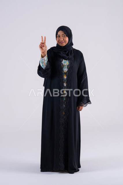 Success and goal achievement: A Saudi woman standing with an expression of joy and happiness. A portrait of a smiling, veiled, Emirati Gulf Arab woman wearing a black abaya, looking at the camera with facial and hand gestures indicating victory and triumph.