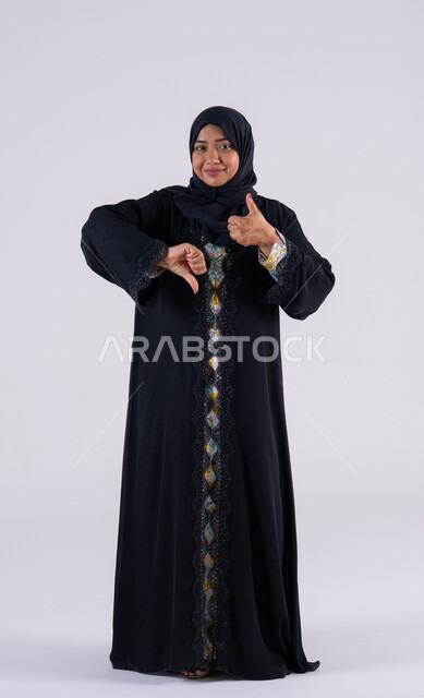 Confusion and hesitation about something. Portrait of an Arab Gulf Emirati woman wearing an abaya and hijab with hand gestures indicating disagreement and approval, dislike and admiration, rejection and acceptance. A Saudi woman giving a thumbs-up and thumbs-down gesture.