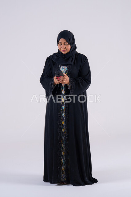 الاطمئنان على الاهل والأصدقاء، سيده سعوديه تستخدم التقنيات والتكنولوجيا الحديثة بالحياة اليومية ،اجراء مكالمه فيديو ،تصفح مواقع وتطبيقات الانترنت، بورتريه لامراه عربيه خليجيه اماراتيه ترتدي العباءه والحجاب تجلس على كرسي ، تمسك الجوال بيدها