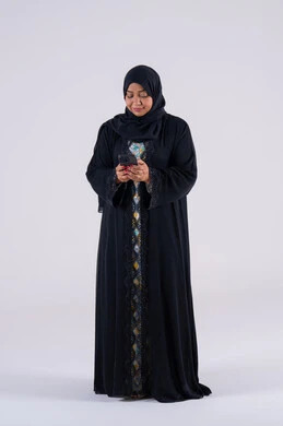 Checking on family and friends, a Saudi woman using modern technology in her daily life, making a video call, browsing websites and applications, a portrait of an Arab Gulf Emirati woman wearing an abaya and hijab sitting on a chair, holding a mobile phone in her hand.