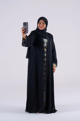 Taking selfies, a Saudi woman using modern technology in daily life, making a video call, chatting with friends, browsing websites and applications, a portrait of an Arab Gulf Emirati woman wearing an abaya and hijab sitting on a chair, holding a mobile phone in her hand.
