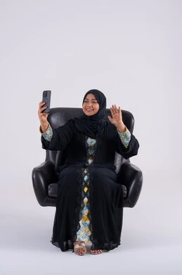 Making a video call, chatting with friends, browsing websites and applications, a portrait of an Arab Gulf Emirati woman wearing an abaya and hijab sitting on a chair, holding a mobile phone in her hand, a Saudi woman using modern technologies in daily life.