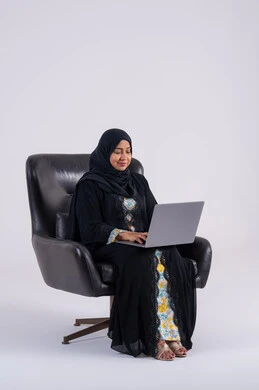 Accomplishing work tasks remotely, using modern technologies in administrative work, portrait of a Saudi Arabian Gulf woman wearing an abaya and hijab sitting on a chair and working on a laptop, making video calls, integrating advanced technology into daily life.