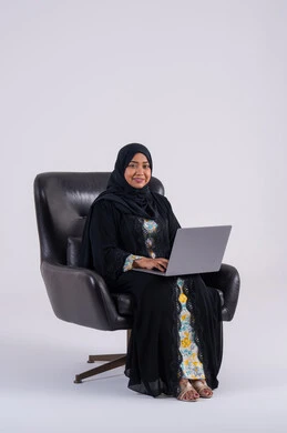 Use modern technologies in administrative work, making video calls, completing work tasks remotely, portrait of a Saudi Arabian Gulf woman wearing an abaya and hijab sitting on a chair and working on a laptop, integrating advanced technology into daily life.