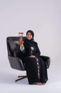 A Saudi woman watches the sand timer, realizing the value of precious moments, the concept of business management and scheduling, a portrait of an Arab Gulf Emirati woman wearing an abaya and hijab, holding a glass hourglass (bankam) in her hand, looking at the camera with gestures of pleasure.