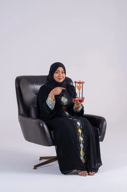 The concept of business management and scheduling, realizing the value of precious moments, a Saudi woman monitoring the sand time, a portrait of an Arab Gulf Emirati woman wearing an abaya and hijab holding a glass sand hourglass (bankam) in her hand, looking at the camera with gestures of pleasure.