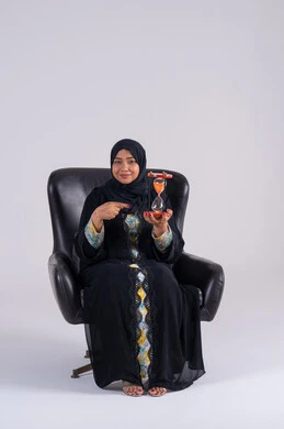 Realizing the value of precious moments, the concept of business management and scheduling, a Saudi woman monitoring the sand time, a portrait of an Arab Gulf Emirati woman wearing an abaya and hijab holding a glass sand hourglass (bankam) in her hand, looking at the camera with gestures of pleasure.