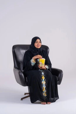 Having a good time at home, gestures of surprise and amazement, home entertainment activities, a Saudi woman eating popcorn, a portrait of an Arab Gulf Emirati woman wearing an abaya and hijab, enjoying watching movies and TV series, and watching TV programs.