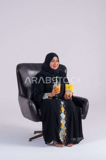 Home entertainment activities, gestures of surprise and amazement, a Saudi woman eating popcorn and cold drinks, a portrait of an Arab Gulf Emirati woman wearing an abaya and hijab, enjoying watching movies and TV series, watching TV programs, and having a good time at home.