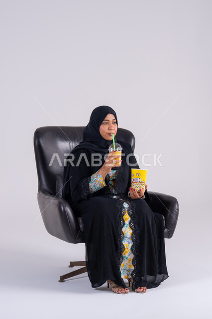 A Saudi woman eating popcorn and cold drinks, home entertainment activities, gestures of surprise and amazement, a portrait of an Arab Gulf Emirati woman wearing an abaya and hijab, enjoying watching movies and TV series, following TV programs, and having a good time at home.
