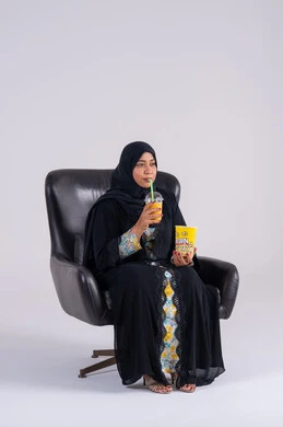 A Saudi woman eating popcorn and cold drinks, home entertainment activities, gestures of surprise and amazement, a portrait of an Arab Gulf Emirati woman wearing an abaya and hijab, enjoying watching movies and TV series, following TV programs, and having a good time at home.