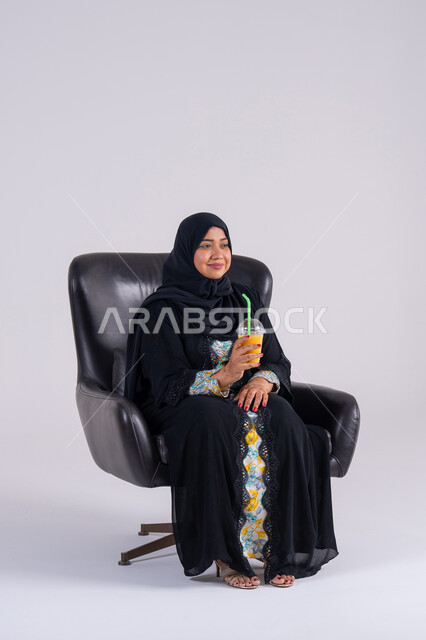 Watching TV shows, having a good time at home, gestures of surprise and amazement, home entertainment activities, a Saudi woman drinking cold drinks, a portrait of an Arab Gulf Emirati woman wearing an abaya and hijab, enjoying watching movies and TV series.