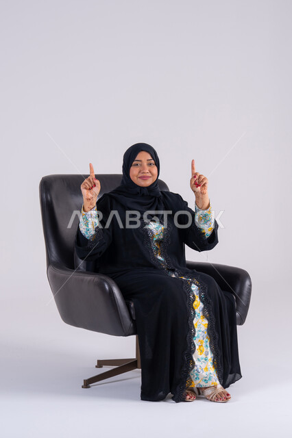 النظر إلى الكاميرا بإيماءات الفرح والسرور،  بورتريه لامراه عربيه خليجيه اماراتيه ترتدي العباءه والحجاب تشير بإصبع السبابة لاعلى إلى شيء ، تعابير تحدد هذا المكان هنا، سيده سعوديه تحدد موقع معين