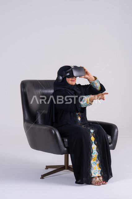 المتعة والتسلية بالمشاهدة عبر نظارة الواقع الافتراضي,  بورتريه لامراه عربيه خليجيه اماراتيه ترتدي العباءه والحجاب تضع نظارات ثلاثية الأبعاد، تنظر بتعابير الاندماج , سيده سعوديه تشير باصبع السبابه الى شئ ما