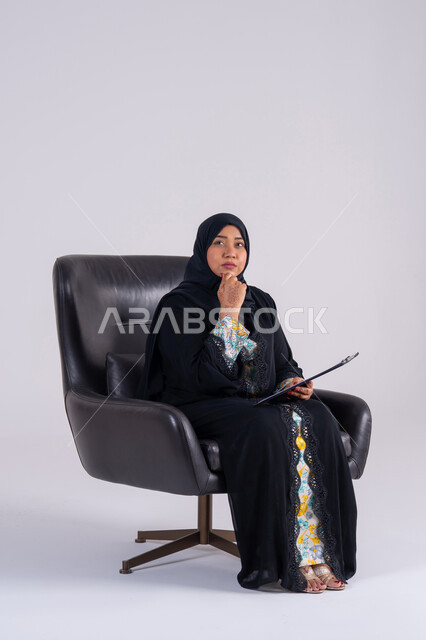 سيده سعوديه تستخدم الاوراق البيضاء الفارغة في العمل، إدارة الأعمال وتنظيمها،  تدوين وتسجيل الملاحظات والمعلومات على الورق, بورتريه لامراه عربيه خليجيه اماراتيه  ترتدي العباءه والحجاب تجلس على كرسي ، وتقوم بكتابة المهام والأهداف اليومية بايماءات التفكير
