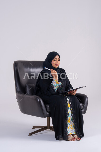 إدارة الأعمال وتنظيمها، سيده سعوديه تستخدم الاوراق البيضاء الفارغة في العمل، تدوين وتسجيل الملاحظات والمعلومات على الورق, بورتريه لامراه عربيه خليجيه اماراتيه  ترتدي العباءه والحجاب تجلس على كرسي ، وتقوم بكتابة المهام والأهداف اليومية