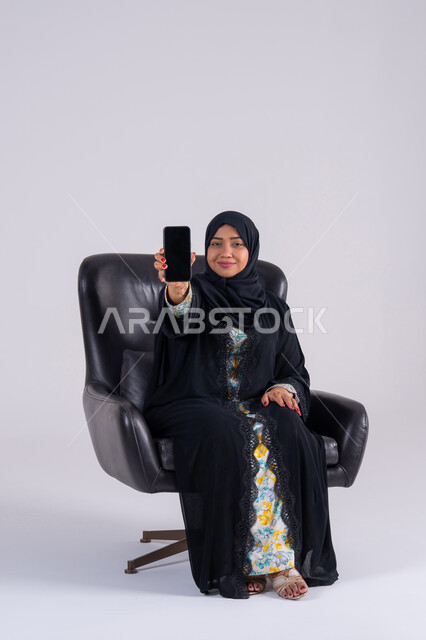 عرض شاشه الهاتف المحمول ، تصفح مواقع وتطبيقات الانترنت،  الدردشة والتحدث مع الأصدقاء، بورتريه لامراه عربيه خليجيه اماراتيه ترتدي العباءه والحجاب تمسك الجوال بيدها، سيده سعوديه تستخدم التقنيات والتكنولوجيا الحديثة بالحياة اليومية