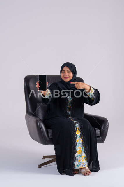 Browsing websites and applications, viewing a mobile phone screen, chatting and talking with friends, a portrait of an Arab Gulf Emirati woman wearing an abaya and hijab holding a mobile phone, a Saudi woman using modern technologies in daily life.