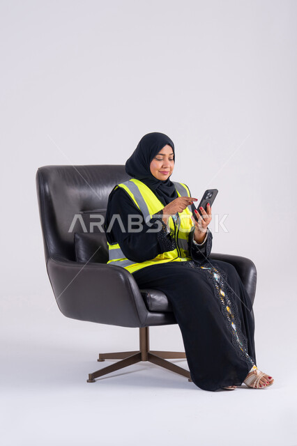 Emirati woman follows up on work and tasks, supervising and monitoring engineering projects, using modern technologies, portrait of a Saudi Arabian Gulf woman wearing an abaya and hijab sitting on a chair and holding a mobile phone in her hand
