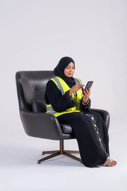 Emirati woman follows up on work and tasks, supervising and monitoring engineering projects, using modern technologies, portrait of a Saudi Arabian Gulf woman wearing an abaya and hijab sitting on a chair and holding a mobile phone in her hand