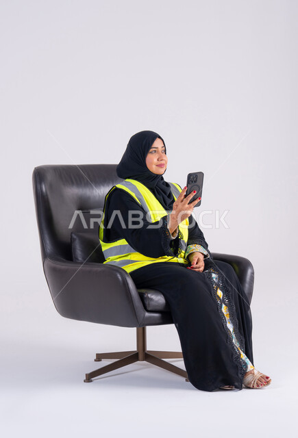 Using modern technology, an Emirati woman follows up on work and tasks, supervising and monitoring engineering projects, a portrait of a Saudi Arabian Gulf woman wearing an abaya and hijab sitting on a chair and holding a mobile phone in her hand