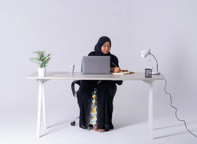 Taking notes and tasks, a Saudi Arabian Gulf woman wearing an abaya and hijab sitting at a desk working on a laptop, drinking hot drinks, using modern technologies in administrative work, an Emirati woman who is focused, dedicated and hard working, employment in offices and companies, integrating advanced technology into daily life