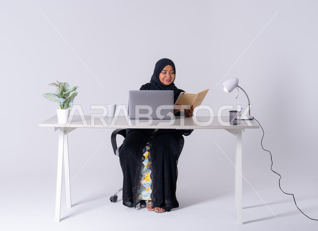 تدوين تقارير العمل, بورتريه لامراه عربيه خليجيه سعوديه ترتدي العباءه والحجاب تجلس على مكتب ،تعمل على جهاز الحاسوب المحمول ، احتساء المشروبات الساخنه، كتابة مهام واهداف العمل ،سيده اماراتيه تقوم باداره الأعمال عن بعد وتنظيمها، استخدم التقنيات الحديثة في الأعمال الإدارية