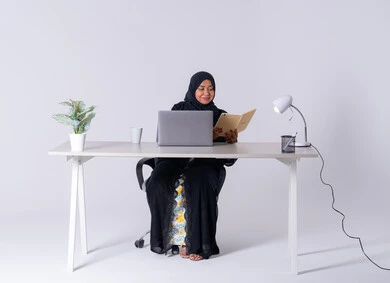 Writing work reports, portrait of a Saudi Arabian Gulf woman wearing an abaya and hijab sitting at a desk, working on a laptop, drinking hot drinks, writing down work tasks and goals, an Emirati woman managing and organizing work remotely, using modern technologies in administrative work