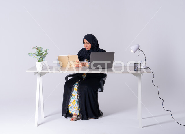 Drinking hot drinks, writing down work tasks and goals, portrait of a Saudi Arabian Gulf woman wearing an abaya and hijab sitting at a desk, working on a laptop, writing work reports, Emirati woman managing and organizing business remotely, using modern technologies in administrative work