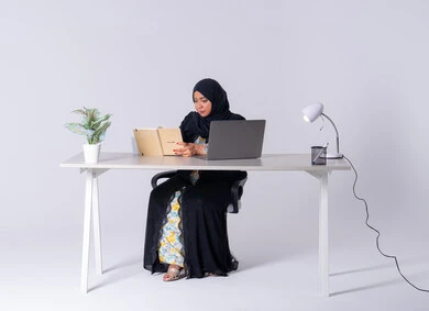Drinking hot drinks, writing down work tasks and goals, portrait of a Saudi Arabian Gulf woman wearing an abaya and hijab sitting at a desk, working on a laptop, writing work reports, Emirati woman managing and organizing business remotely, using modern technologies in administrative work