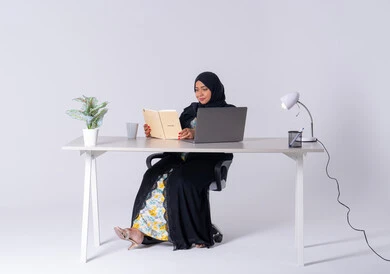 Writing work tasks and goals, drinking hot drinks, portrait of a Saudi Arabian Gulf woman wearing an abaya and hijab sitting at a desk, working on a laptop, writing work reports, an Emirati woman managing and organizing business remotely, using modern technologies in administrative work