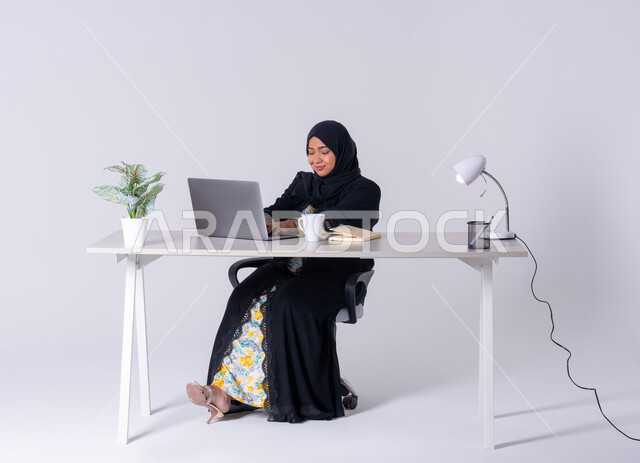 Focus, dedication and hard work, employment in offices and companies, use modern technologies in administrative work, portrait of a Saudi Arabian Gulf woman wearing an abaya and hijab sitting at a desk and working on a laptop, integration of advanced technology into daily life, an Emirati woman drinking hot drinks