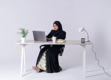 Focus, dedication and hard work, employment in offices and companies, use modern technologies in administrative work, portrait of a Saudi Arabian Gulf woman wearing an abaya and hijab sitting at a desk and working on a laptop, integration of advanced technology into daily life, an Emirati woman drinking hot drinks