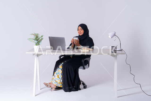 بورتريه لامراه عربيه خليجيه سعوديه ترتدي العباءه والحجاب تجلس على مكتب وتعمل على جهاز الحاسب المحمول ، التركيز والتفاني والعمل الجاد،التوظيف في المكاتب والشركات ،استخدم التقنيات الحديثة في الأعمال الإدارية ، دمج التكنولوجيا المتطوره فى الحياه اليوميه ، سيده اماراتيه تقوم باحتساء المشروبات الساخنه