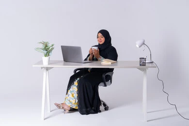 Portrait of a Saudi Arabian Gulf woman wearing an abaya and hijab sitting at a desk working on a laptop, focus, dedication and hard work, employment in offices and companies, use of modern technologies in administrative work, integration of advanced technology into daily life, an Emirati woman drinking hot drinks