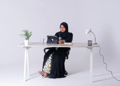 Use modern technologies in administrative work, drinking hot drinks, writing down work tasks and goals, taking notes and recording information on paper, an Emirati woman managing and organizing business remotely, a portrait of a Saudi Arabian Gulf woman wearing an abaya and hijab sitting at a desk, working on a tablet