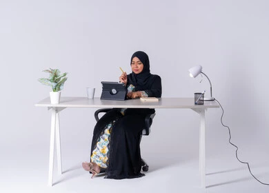 Drinking hot drinks, writing down work tasks and goals, taking notes and recording information on paper, an Emirati woman managing and organizing business remotely, a portrait of a Saudi Arabian Gulf woman wearing an abaya and hijab sitting at a desk, working on a tablet, using modern technologies in administrative work