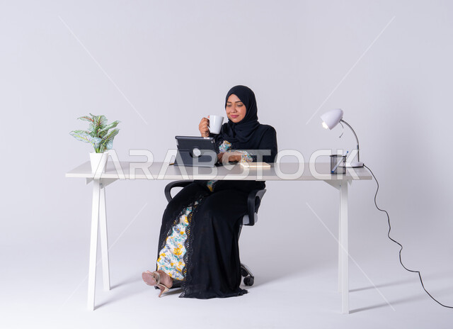 Writing down work tasks and goals, drinking hot drinks, taking notes and recording information on paper, an Emirati woman managing and organizing business remotely, a portrait of a Saudi Arabian Gulf woman wearing an abaya and hijab sitting at a desk, working on a tablet, using modern technologies in administrative work